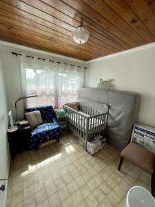 A bedroom with a wooden ceiling, a big double window, a crib, chairs, and a mattress leaning up against the wall.